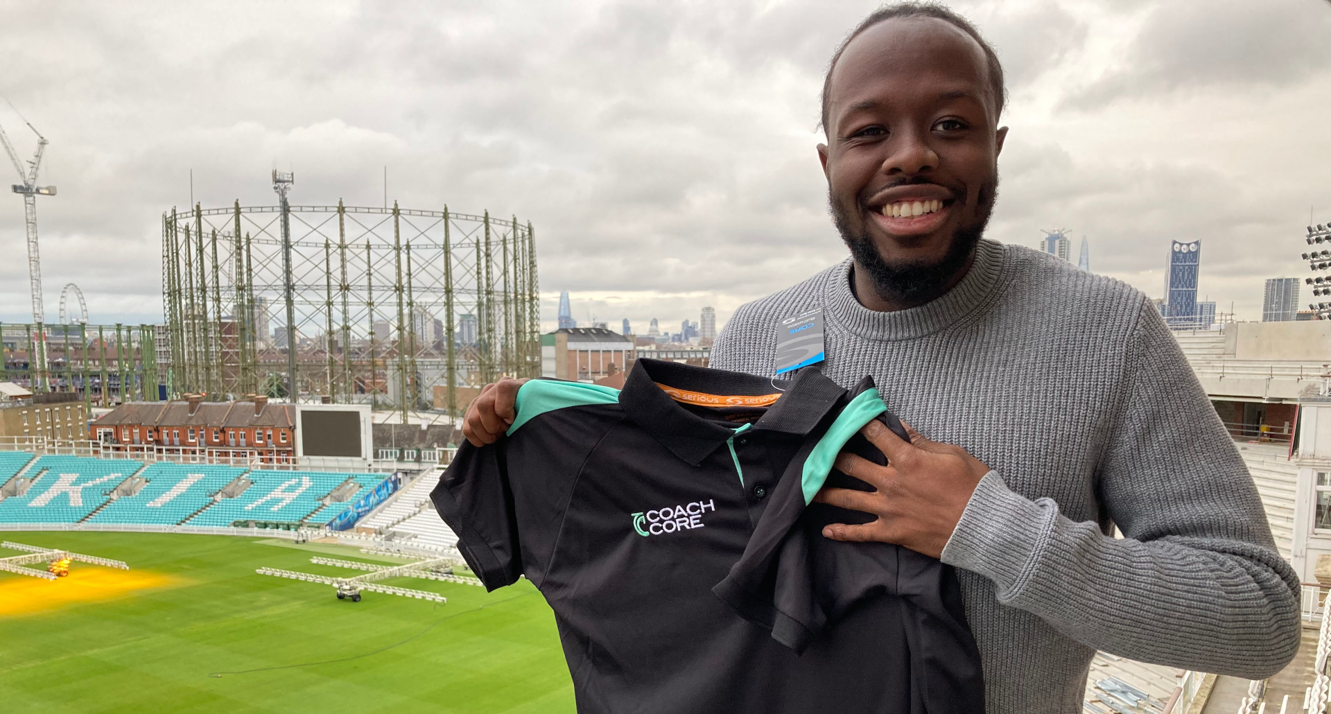 Terrell stands on balcony above cricket pitch, holding Coach Core shirt