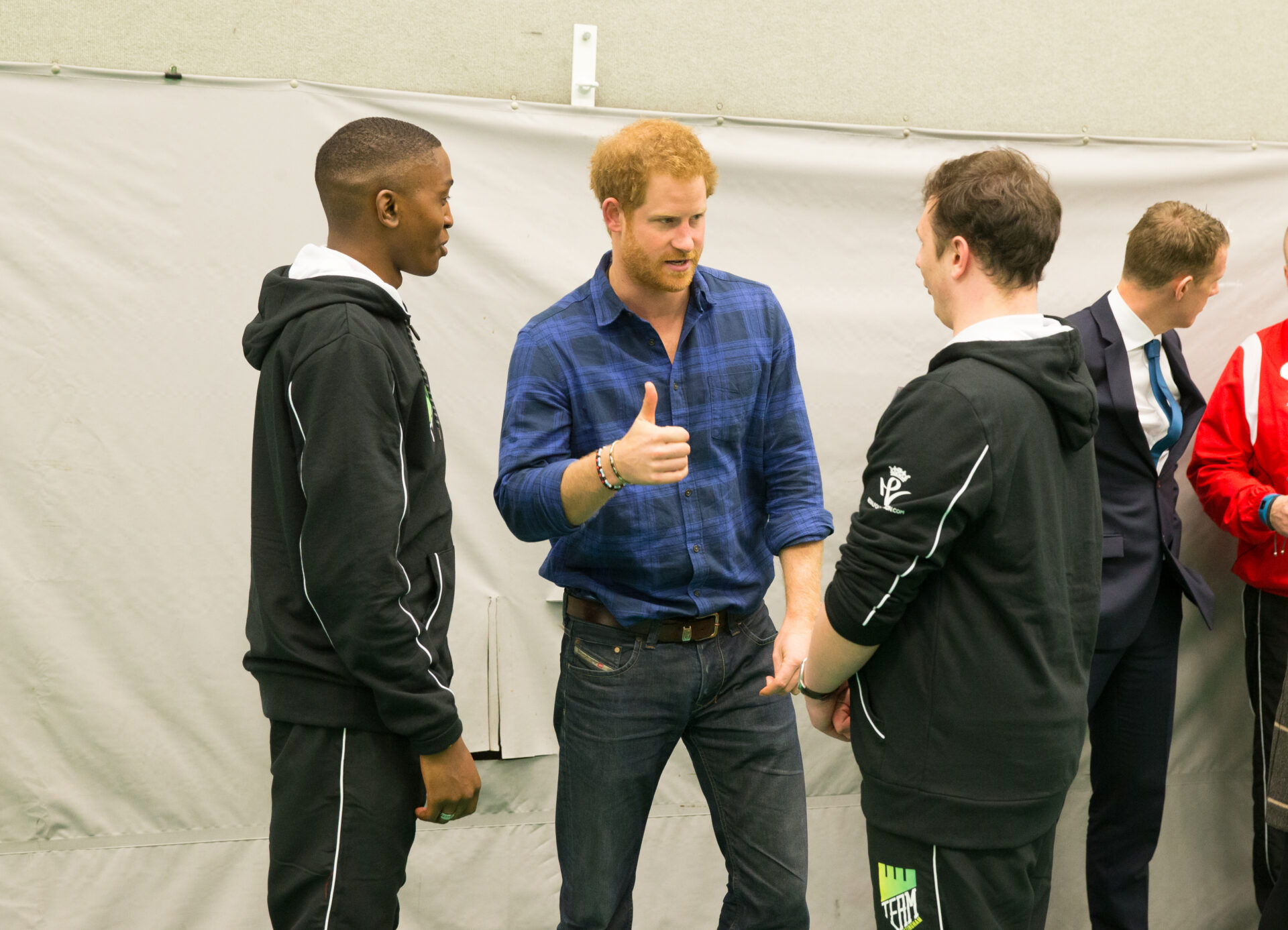 3 men stand in a group. The central man - Prince Harry - gives a thumbs up to one of the others